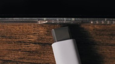 Yukarıdan aşağıya USB-C kablosu akıllı telefon portuna bağlanıyor. Cihaz açık bir bavula ve ahşap bir masa arkasında duran siyah bir ekrana sahip. Yükleme veya veri transferi için bir aygıt bağlanıyor.