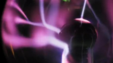 Pembe ve mor ışık akımları yayan merkezi elektrotlu bir plazma lambasının yan açısı. Parlak boşalımlar cam hazne boyunca kıvrılıp akıyor. Koyu bir arkaplana karşı akışkan hareket.