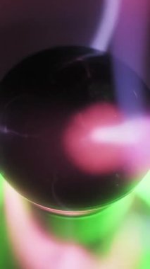 Merkezi bir elektrottan hareket eden pembe ve mor elektrikli cıvatalı bir plazma küresinin dikey yakın çekimi. Parlak yeşil bir tepenin üzerinde iyonize gaz filizleri parlıyor. Koyu soyut arkaplan.