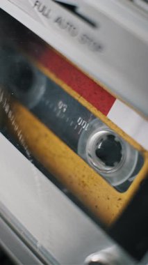 Sarı bir ses kasetinin dikey, açılı makro görüntüsü. Etiketinde numaralar olan özet, retro teyp kayıt cihazının gümüş güvertesinde yavaşça dönüyor..