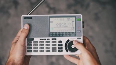 Beyaz portatif bir radyodaki FM istasyonlarını aktif olarak arayan kişinin perspektifi, arka plandaki LCD ekranda hızla değişen rakamlarla büyük bir elle ayarlama kadranı kullanıyor..