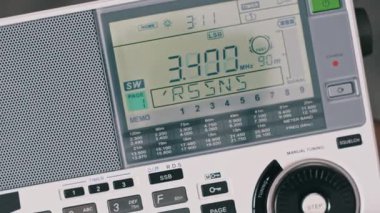 Taşınabilir bir kısa dalga radyosunu ayarlayan birinin yukarıdan aşağı görüntüsü. Bir parmak, dijital istasyon bilgisi ve sinyal gücünü gösteren LCD ekranındaki SW frekansını değiştiren elle arama ayarlarını yapar.