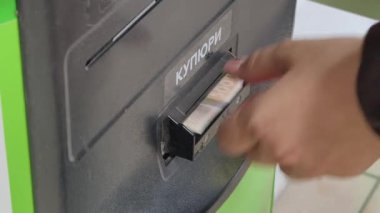 Ödeme terminalinden bir fatura alıcısına banknot sokan birinin yakın çekimi. Siyah makinenin para aktarımı için Kiril alfabesi metni var.