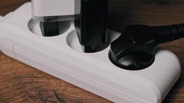 Yakından bir erkek eli, beyaz plastik bir şeritten siyah bir güç kablosu ve beyaz bir adaptör çıkarıyor. Tahta masadaki dizilim, cihazları güçten ayırarak enerji korumasını gösterir.