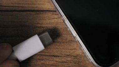 Modern bir akıllı telefon portuna beyaz bir USB-C konnektörü yerleştiren parmaklara aşırı yakın çekim. Ahşap bir masanın üzerindeki şeffaf bir çantadaki cihaz enerji transferini temsil eden pilini şarj eder..