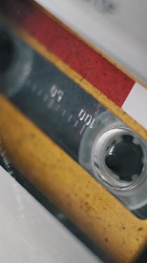 Güvertede çalan eski sarı ve kırmızı bir kasetin dikey, makro görüntüsü. Teyp, etiketteki zaman işaretçilerini geçmişteki analog ses ortamını oynatırken görüntüler..