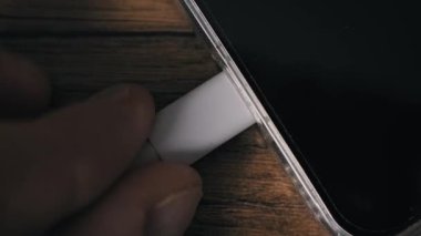 Beyaz bir USB-C güç kablosunu akıllı telefondan söken bir elin açılı yakın çekimi. Şeffaf bir koruyucu çantanın içindeki cihaz ahşap bir yüzeye oturmuş ve mobil cihazın elektriğini kesiyor..