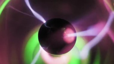 Merkezi elektrotun etrafında dönen makro görüntülü plazma topları. Pembe ve yeşil iyonize gaz filizleri şeffaf kürenin içinde hareket eder. Karanlık bir arkaplanda bulanık ışık halkaları.