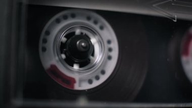 Güvertede çalan şeffaf bir ses kasetinin makro görüntüsü, beyaz plastik bir makara hızla dönüyor. Karanlık manyetik bant gevşer ve karanlık iç kısımdaki kırmızı lider bandını ortaya çıkarır..