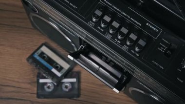 Üst kattan bakıldığında, sarı etiketli eski bir ses kasedini güverteyi kapatan eski bir teybe yerleştirirken ve arka plandaki ahşap masada görülebilen diğer karışık kasetlerle oynat tuşuna basarken görülüyor.