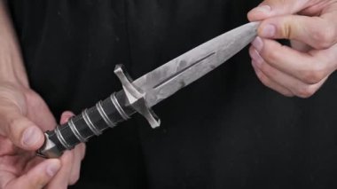 El ele tutuşun ve küçük süslü bir hançeri dikkatlice inceleyin. Eski silahta ayrıntılı bir kabza ve kaba elle dövülmüş metalik bir bıçak var. Koyu arkaplan.