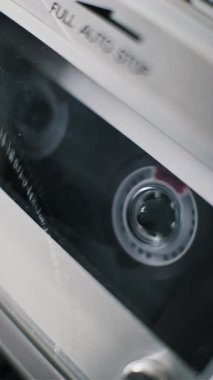 Eski bir kayıt cihazında çalan şeffaf bir ses kasetinin dikey ve yakın çekimi. Tedarik makarası, manyetik bant eski analog elektronik hi-fi cihazının içinde açılırken dönüyor..