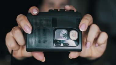 Fransız manikürlü bir kadın siyah taşınabilir bir kaset kaydedici taşıyor. İç bant makaraları dönüyor, arka planda bulanık yüzü olan kaydı ya da kaydı gösteriyor.