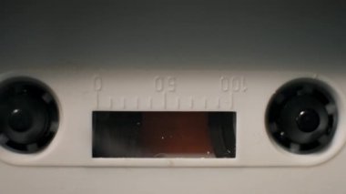 Eski beyaz kaset çalarken çekilen statik makro çekim. Siyah makaralar sürekli döner, manyetik filmi şeffaf pencereden geçirir ve plastik kabukta ölçüm işaretleri bırakır..