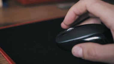 Yakından bir erkek eli kırmızı kenarlı bir fare üzerinde siyah bir bilgisayar faresi kullanıyor. Parmak kaydırma tekerleği ve sol düğmeye tıklayarak ahşap masadaki internet gezintisi ve gezinti için tıklıyor.
