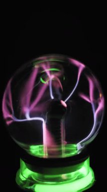 Merkezi elektrottan gelen açılı plazma küre pembe yeşil cıvatalar. İyonize gaz filizli cam küre, arka planda elektrik akımlarını hareket ettiriyor. Yeşil ışıklandırılmış plastik taban. Yüksek voltajlı çekim.