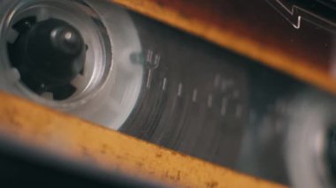 Eskimiş sarı bir teypte çalan eski bir ses kasedinin açılı makro görüntüsü. Manyetik film, retro bir kurulumda destek makarasında gevşiyor..