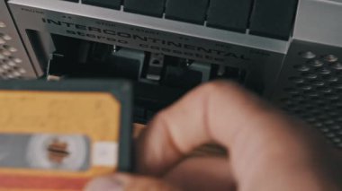 Eller yukarı aşağı görünümlü, eski bir ses kasedini sarı etiketli eski bir stereo çalar içine yerleştiriyor, güverteyi kapatıyor ve gümüş makinedeki çalma tuşuna basıyor..