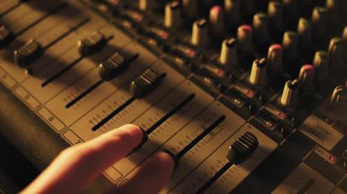 Canlı müzik etkinliği sırasında mavi ve mor sahne ışıklandırması altında desibel işaretlerini ve kontrol düğmelerini gösteren profesyonel bir ses panosundaki birden fazla fadoru kaydıran parmakların ayrıntılı görünümü.
