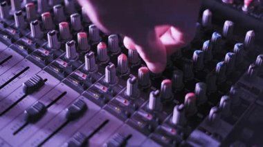 Ses mühendisi elinin yan açısı, ses karıştırma konsolundaki çoklu faderleri ayarlıyor. Mor ve mavi ışıkların altında yanıp sönen düğmeler ve desibel işaretleyicileri ile tam kanal kontrolünü gösteriyor..