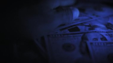 Mavi ışığın altındaki karanlık bir odada büyük bir yığın 100 dolarlık banknotu sayarken makro görüntüyü kapat. Hızlı hareket zenginlik, ödeme veya yasadışı mali işlemler temaları vurguluyor.