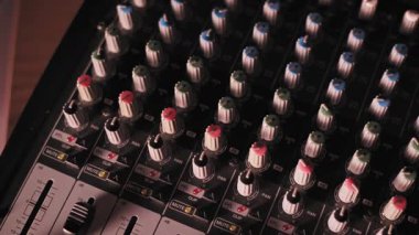 Profesyonel ses karıştırma konsolunun yüksek açılı tavası düğme ve kontrol düğmelerini gösteriyor. Stüdyonun ışıklandırması dinamik bir şekilde ılık turuncudan havalı mor renklendirme ekipmanlarına kayar..