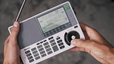 LCD ekranında FM frekansı değişirken, erkek ellerinin beyaz, taşınabilir, antenli bir radyo alıcısını tutarken elle ayarlama düğmesini döndüren bir başparmağın açılışı...