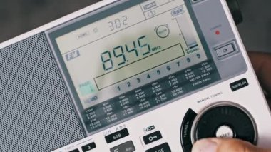 Bir insan, büyük bir düğmesi olan beyaz taşınabilir çoklu bantlı bir radyoyu elle ayarlar. Detaylı LCD ekranına yakın çekim FM frekans sayısının arttığını gösteriyor..