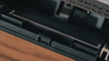 Eski bir ses kasedini, üzerinde sarı etiket olan eski bir gümüş teyp teybine yerleştiren bir elin yakın çekimi. Mekanizma bandı kabul eder ve ahşap bir yüzeye kapatır..