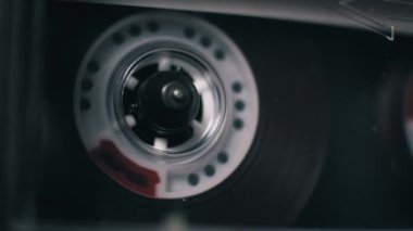 Bir çalgıcıda çalan şeffaf bir ses kasetinin yakın plan görüntüsü. Beyaz makaralar, 90 'ların nostaljisini çağrıştıran, karanlık bir ortamda sürekli dönen, kıvrımlı manyetik film..