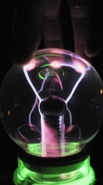 Cam küreye dokunan bir elin dikey yakın çekimi. Beyaz ve pembe plazma şeritleri parmakları takip eder. İyonize gaz filizleri yeşil bir zemin üzerinde parıldıyor. Etkileşimli bilim aygıtı.