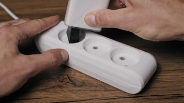 Beyaz bir adaptör, siyah bir şarj aleti ve siyah kabloyu ahşap bir masanın üzerindeki beyaz bir uzatma bloğuna takan ellerin yan açısı. Çoklu aygıtlar ve ev aletleri çalıştırılıyor ve bağlanıyor.