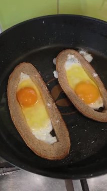 Yuvarlak kahverengi ekmek dilimlerinin içinde pişen iki yumurtanın yukarıdan görünüşü. Siyah, yapışmaz bir tavada cızırdayan yağla birlikte. Turuncu sarılar ve pişmiş beyazlar. Basit kahvaltı tarifi ve ev yapımı yemek hazırlama..