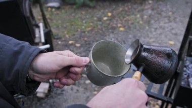 Eller, bakır bir kafesten, açık havada kullanılan eski bir diş minesi bardağına, dumanı tüten koyu kahve döker. Sıcak içecek, doğadaki bir kamp gezisi sırasında taze olarak servis edilir..