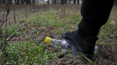 Siyah bir spor ayakkabı giyen bir erkek ayağı çimlerin üzerinde atılmış plastik bir şişeye sıkıca basar. Eylem, atıkların ezildiğini ve yeşil alanlardaki kirliliğin yaygınlığını gösteriyor..