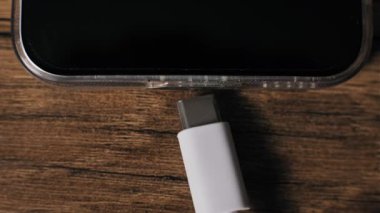 Beyaz bir USB-C kablosunun yukarıdan aşağı makro görüntüsü net bir koruyucu çantadaki akıllı telefona bağlanıyor. Modern cihaz, bir güç kaynağına bağlanan kahverengi ahşap bir masanın üzerinde duruyor..