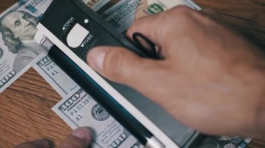 Bir erkeğin portatif bir sahte kimlik tarayıcısını 100 dolarlık banknotlar üzerinde hareket ettirdiği yakın plan görüntüsü. Süreç mali doğrulama ve para birimi denetimini gösteriyor.