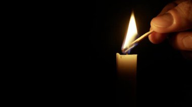 El, siyah arka planda yanan bir kibrit çöpüyle tek bir mum yakar. Issız alev, karanlığın içinde sıcak, samimi bir parıltı sağlar..