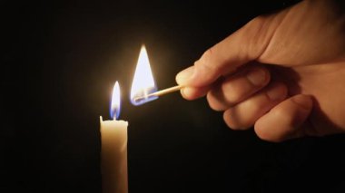 El, beyaz bir mum fitiline bir kibrit tutar ve karanlık bir odada tek bir alev yakar. Ilık, izole ışık saf siyah bir arkaplana karşı titreyerek, ciddi ve barışçıl bir ruh hali yaratır..