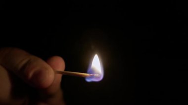 Ateş odunu yavaş yavaş yakarken parmaklar tek bir kibrit çöpünü tutuyor. Makro yakın çekim siyah bir arkaplanda izole edilir ve tek ışık kaynağı alevdir..