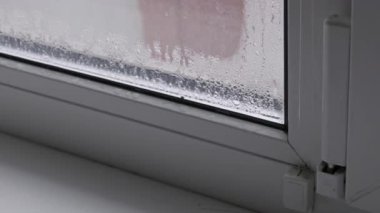 Soğuk cam pencere camında su damlacıklarıyla yoğun bir yoğunlaşma var. Kamera yavaşça yakınlaşıyor, soğuk kış havasında yüksek içerideki nem oranını gösteriyor..