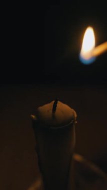 Yanan kibrit çöpü, karanlık bir odada beyaz bir mumun fitilini yakar. Sıcak, titreyen alev, siyah arka planda samimi ve umut dolu bir parıltı yaratır..