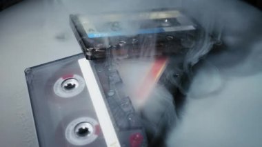 Üç klasik ses kaseti karanlık ve yansıtıcı bir yüzeyde duruyor. Kalın beyaz duman yavaş yavaş dağılır, karamsar ve dramatik bir çekimle retro medyayı ortaya çıkarır..