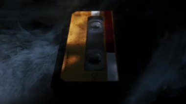 Üzerinde sarı etiket bulunan klasik ses kaseti koyu renkli bir yansıtıcı yüzeyde duruyor. Esrarengiz beyaz duman dalgalanır ve girdaplar, eski nesneyi gizler..