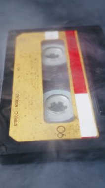 Sarı etiketli klasik ses kaseti karanlık bir yüzeyde yavaşça ortaya çıkar. Kalın beyaz duman dalgalanır, gizemli ve nostaljik bir atmosfer yaratır..