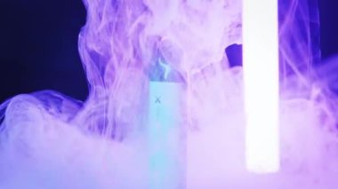 Tek kullanımlık elektronik sigara siyah arka planda duruyor. Etrafında yoğun bir buhar dalgası var. Renkli neon lambalar, mor, mavi ve yeşil tonlarda dönen dumanı aydınlatır..