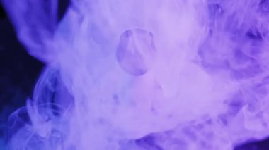 Tek kullanımlık beyaz sigara, karanlık bir yüzeyde döner. Mor ve mavi ışık altında cihazın etrafında yoğun buhar bulutları uçuşuyor..