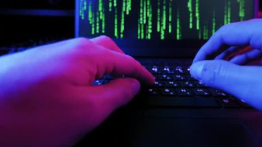 Bilgisayar korsanlarının elleri ekranında yeşil kod olan bir dizüstü bilgisayarda hızla yazarlar. Karanlık oda dramatik mavi ve mor neon ışıkla aydınlanır..
