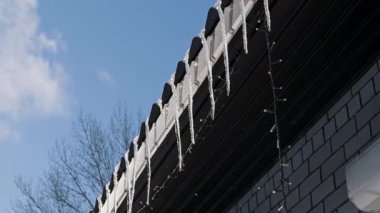 Buz saçakları, mavi gökyüzüne karşı karanlık bir çatı oluğunda asılı. Kamera yavaşça uzaklaşırken parlak kış güneşi buzun üzerinde parlıyor..