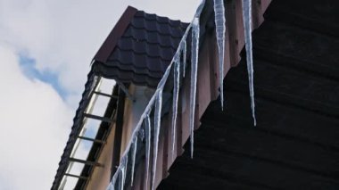 Buz saçakları bulutlu bir gökyüzüne karşı karanlık bir çatının kenarından sarkıyor. Düşük açılı çekim soğuk, kışın serin atmosferini yakalar.